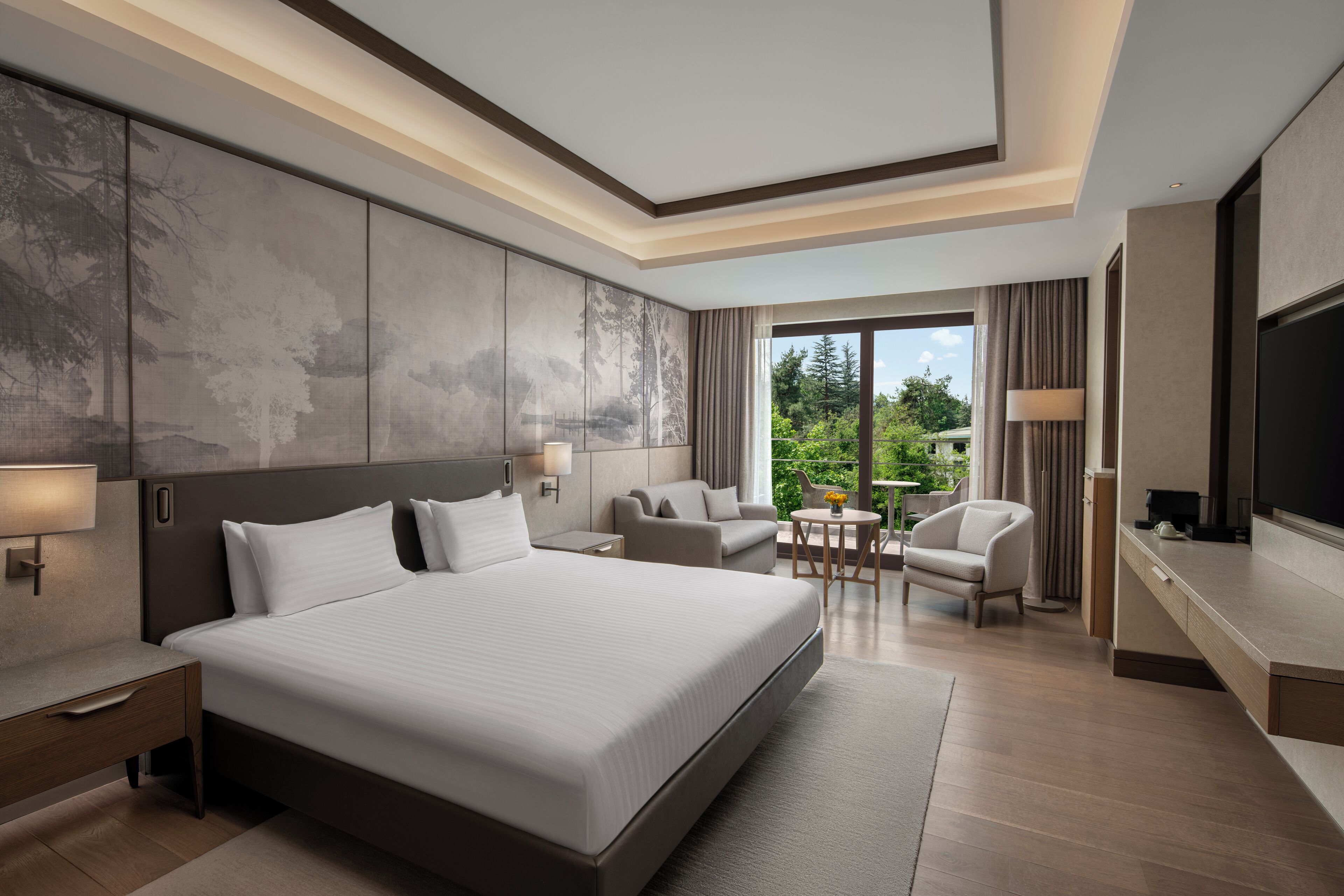 Une chambre d’hôtel moderne dotée d’un grand lit, d’une télévision, d’un bureau et d’un balcon offrant une vue sur la verdure.