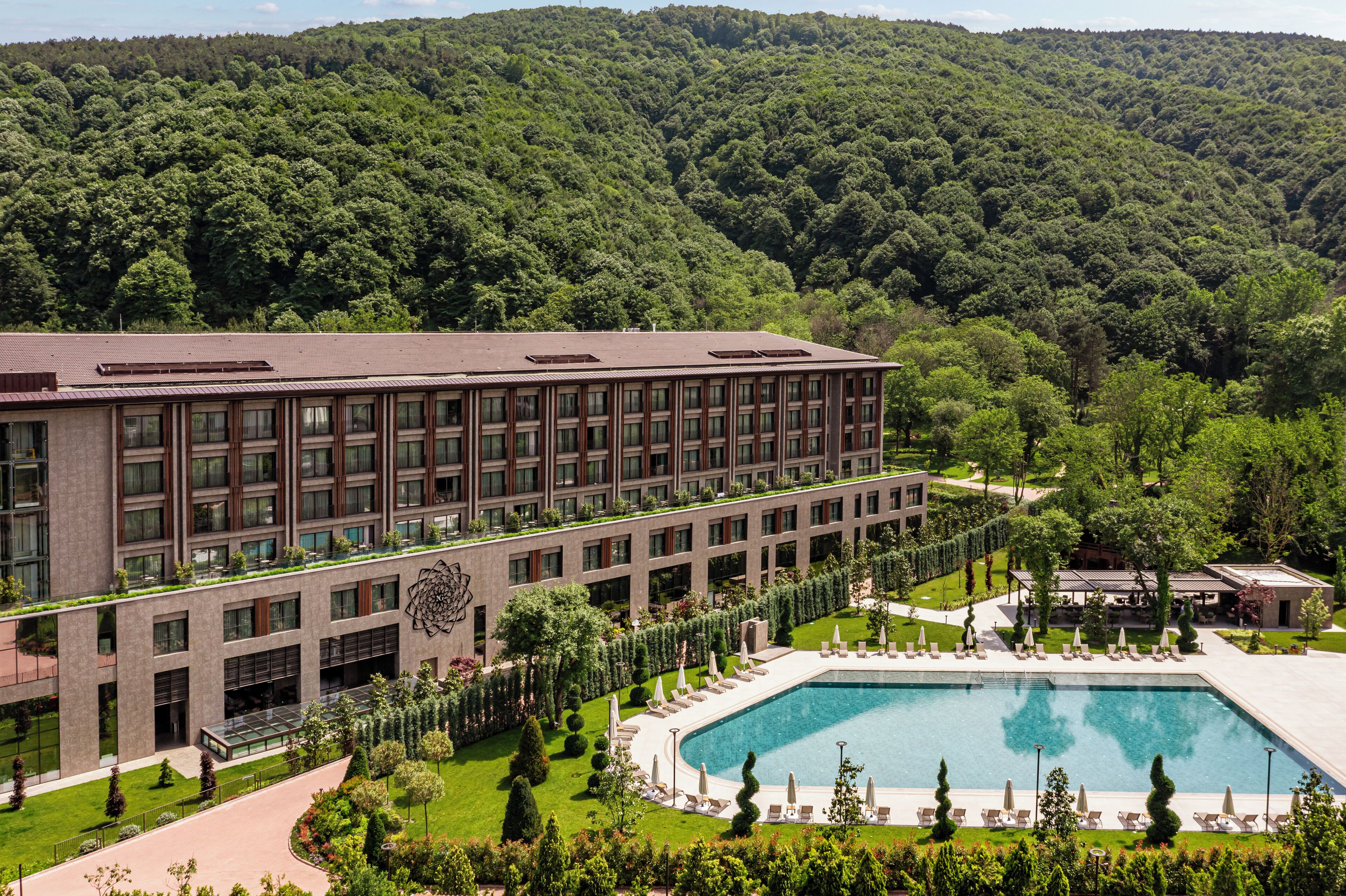 A modern hotel with a large pool, surrounded by lush greenery and a forested hillside.