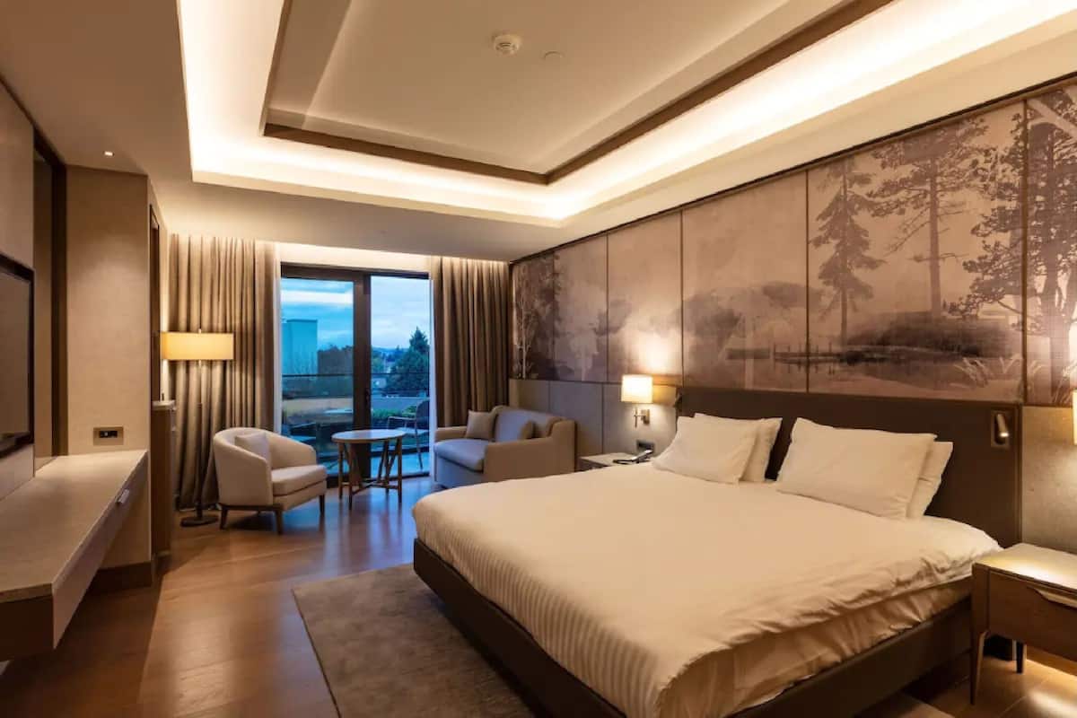 Ein Hotelzimmer mit einem großen Bett, einem Sofa, einem Sessel, einem Schreibtisch und Blick ins Freie.