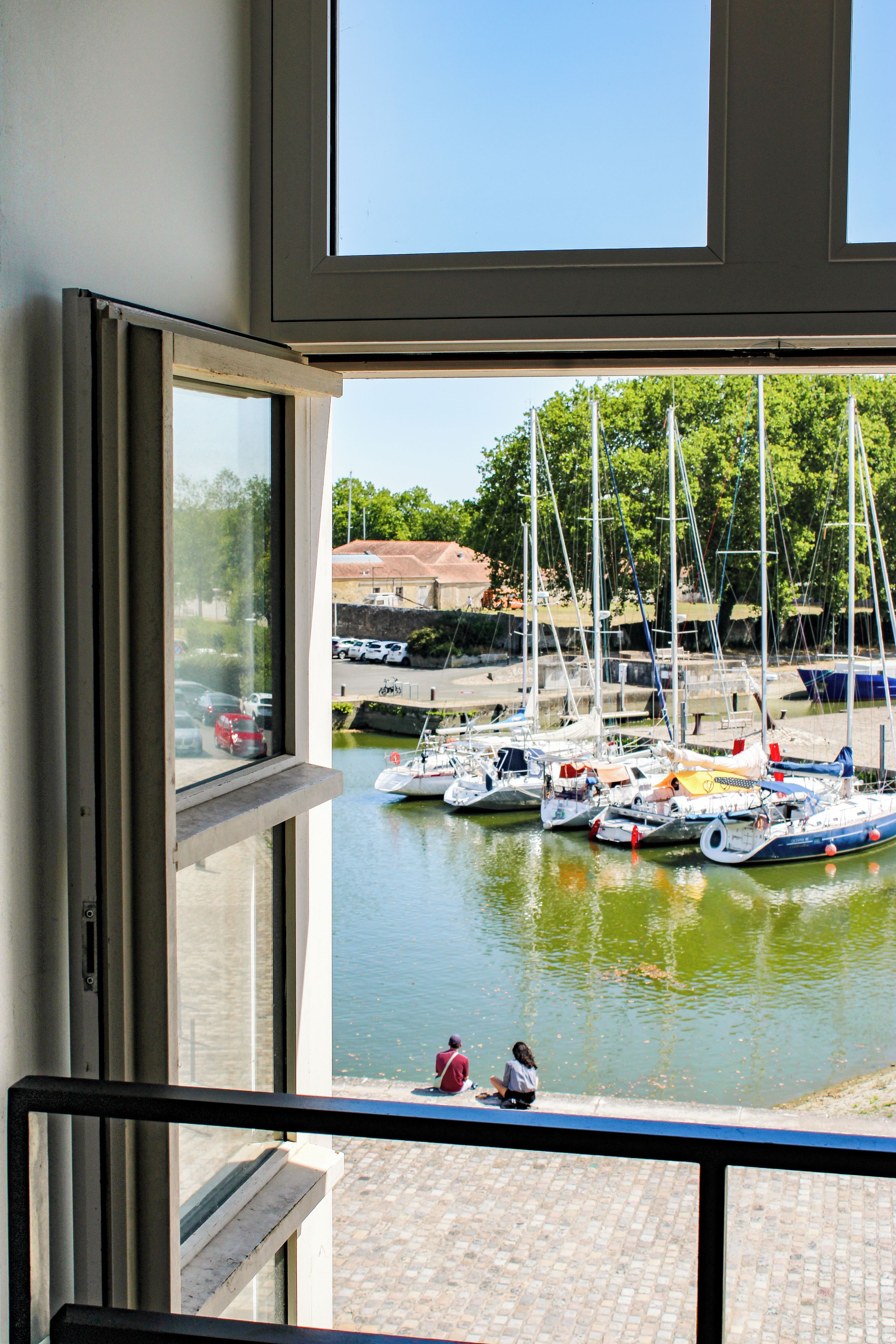 Vista desde una ventana con vista a un puerto deportivo con barcos amarrados, un paseo marítimo pavimentado y un edificio al fondo.