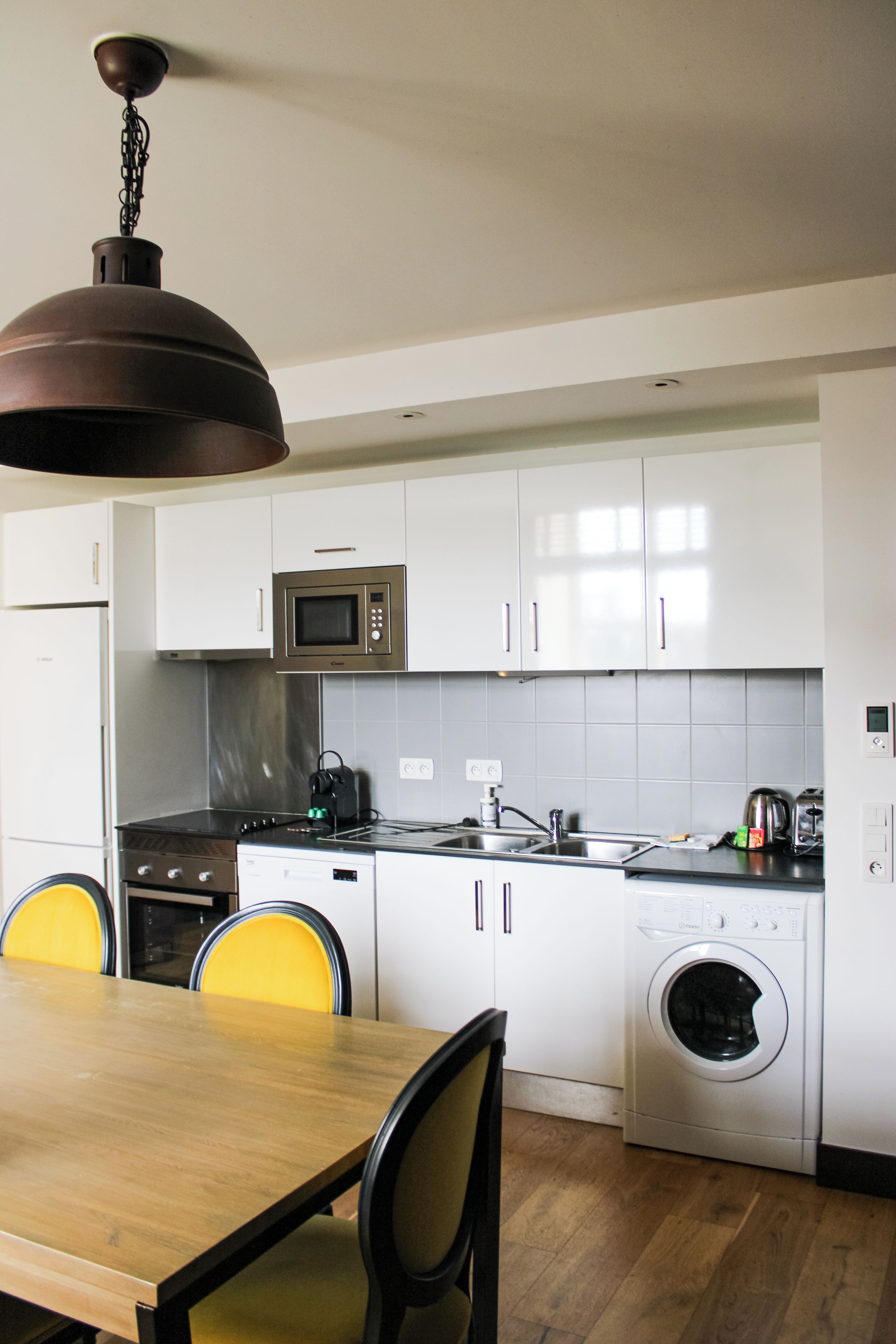 A modern kitchen with white cabinets, a microwave, a stove, a sink, and a washing machine. There is a wooden dining table with chairs.