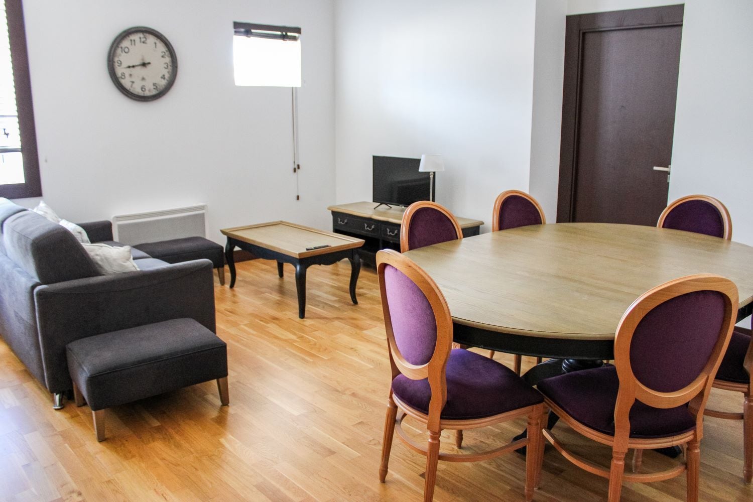 Una habitación con una mesa redonda de madera, sillas tapizadas en color morado, un sofá, una pequeña mesa de centro de madera, un televisor y un reloj en la pared.