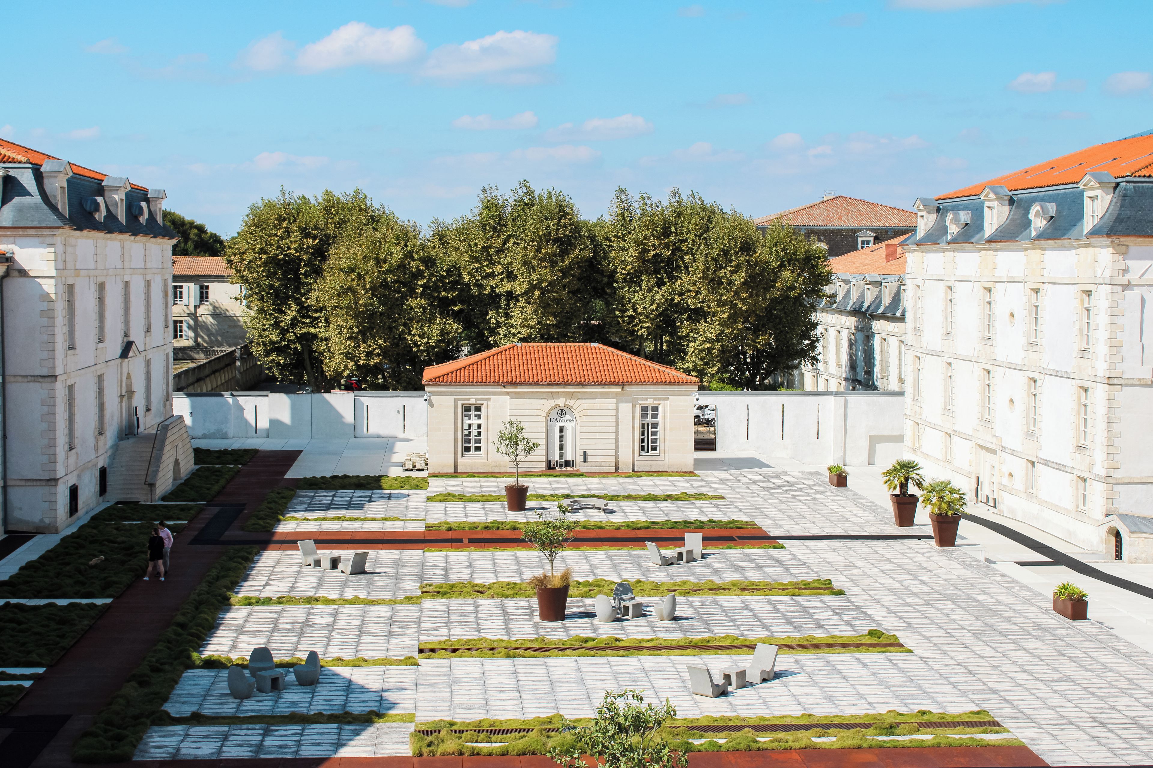 Une cour intérieure avec un bâtiment central, des espaces aménagés pour s’asseoir et un décor arboré.