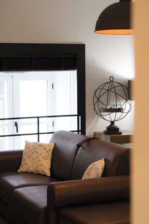 Ein braunes Ledersofa mit gemusterten Kissen, ein Fenster mit schwarzem Rahmen und eine dekorative Metallkugel auf einem Tisch.