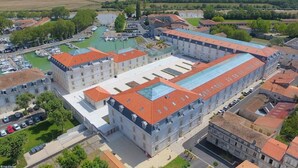 Un edificio grande e histórico con varias alas y un patio central, rodeado por un puerto deportivo y campos verdes.