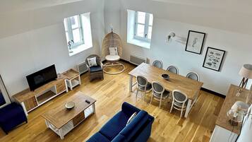 A spacious living room with a wooden floor, a blue sofa, a dining table with chairs, a television, and a wicker chair.