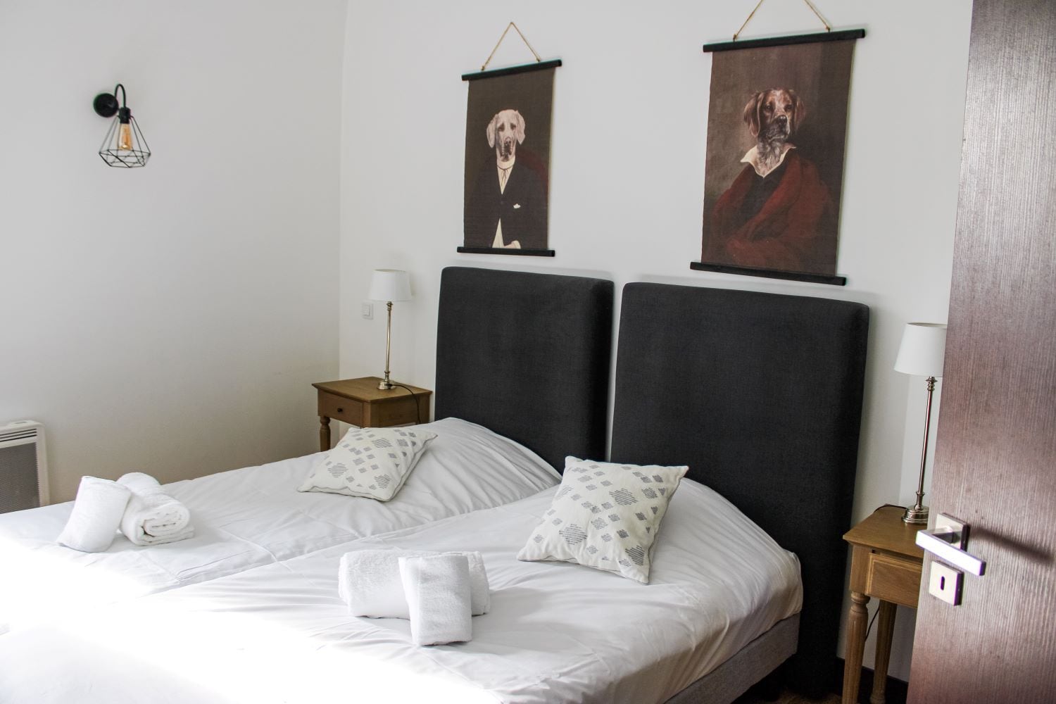 A hotel room with a bed, two pillows, a nightstand with a lamp, and two wall-mounted pictures.