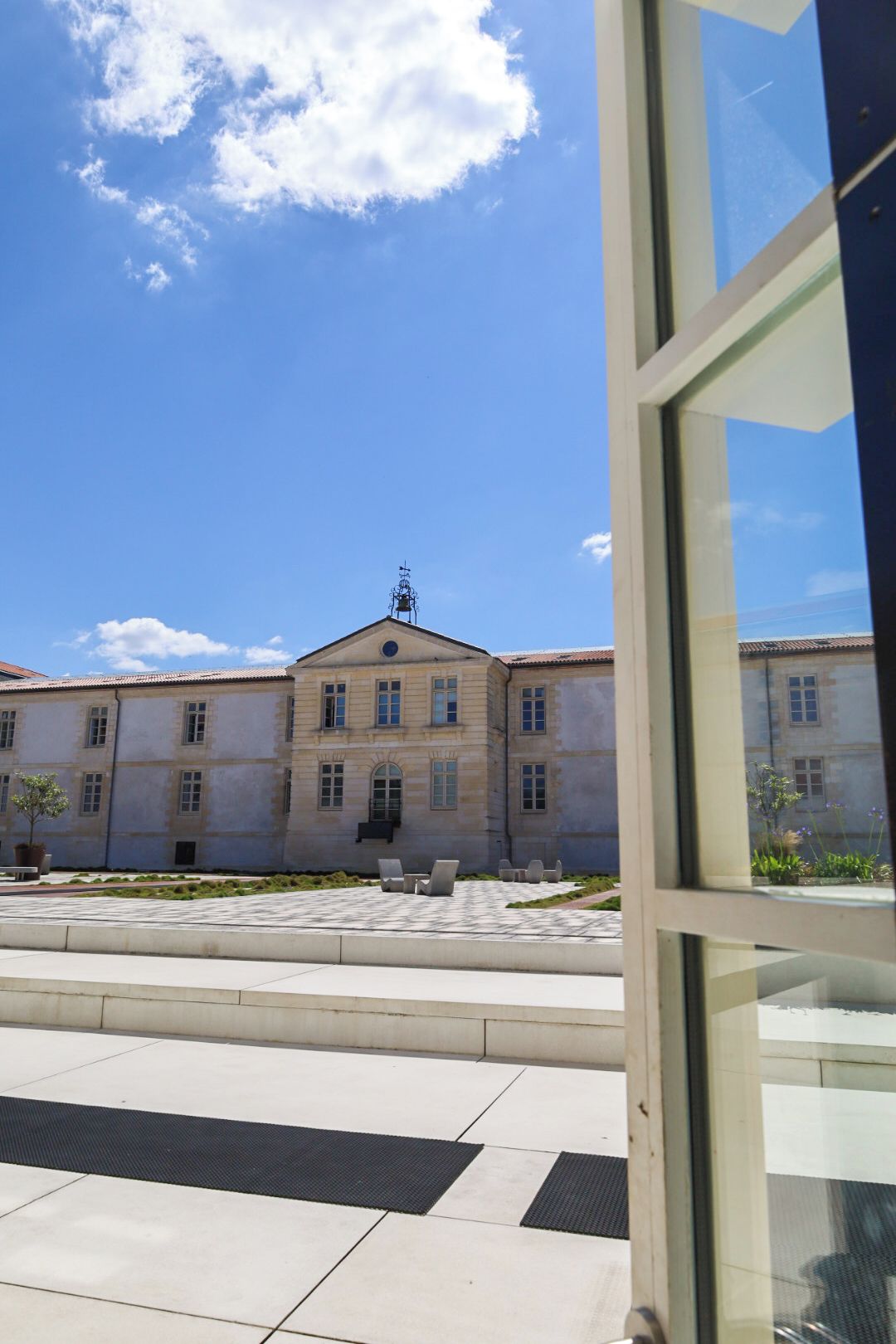 A building with a central dome and multiple windows, a paved courtyard with seating, and a clear sky.