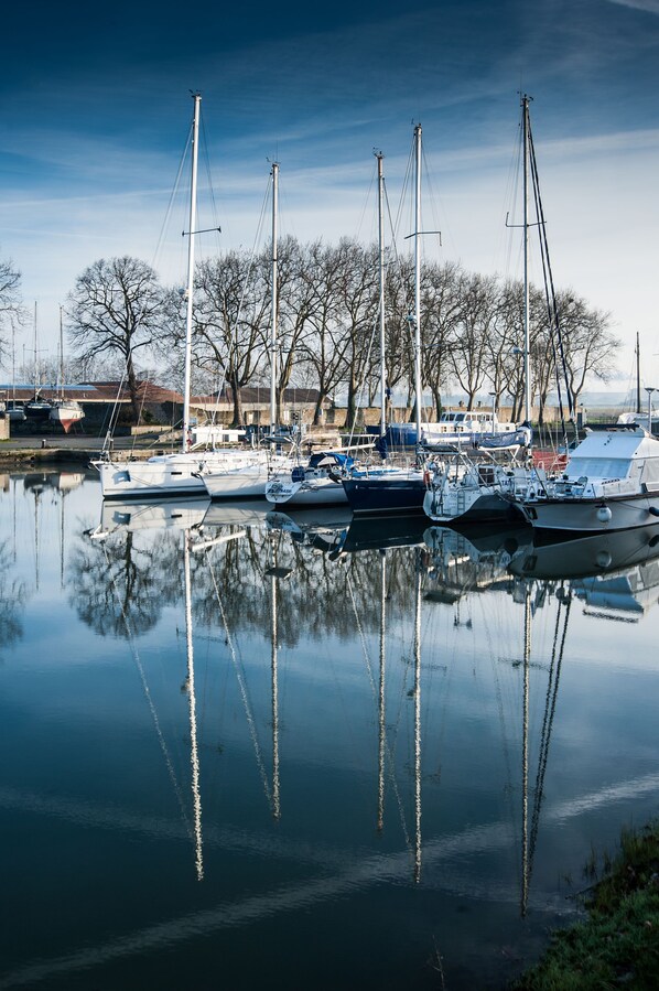 Un port de plaisance où sont amarrés plusieurs voiliers, une eau calme qui reflète les bateaux et les arbres, et un ciel dégagé.
