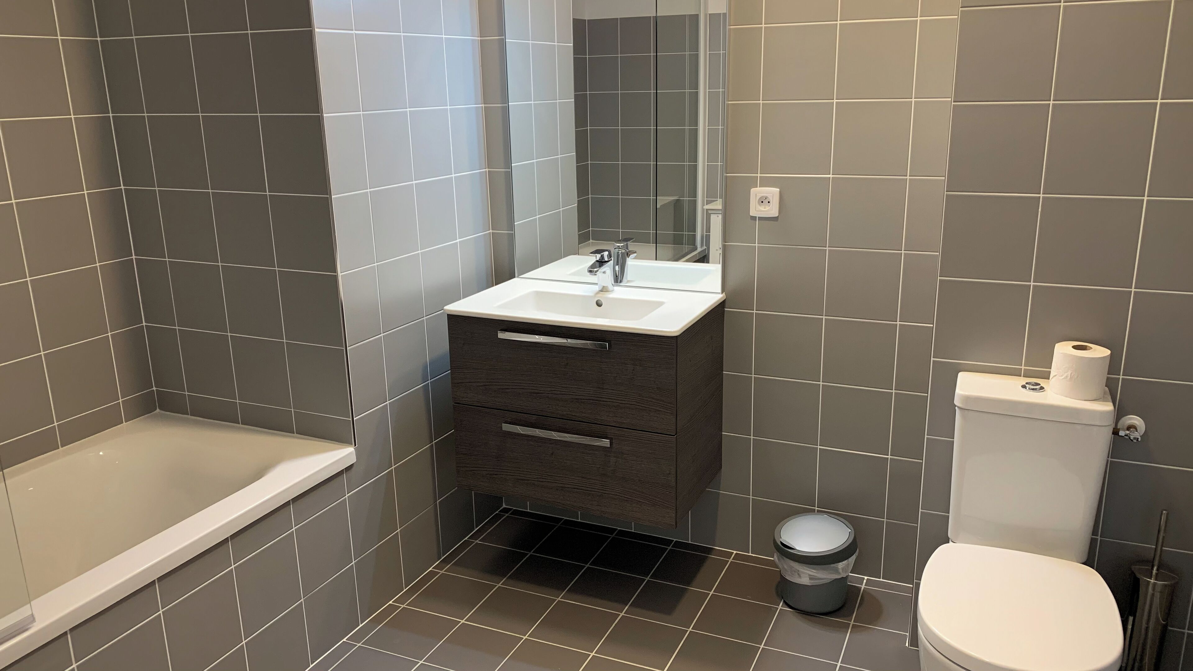 A bathroom with a bathtub, a sink with a mirror, a toilet, and a trash bin.