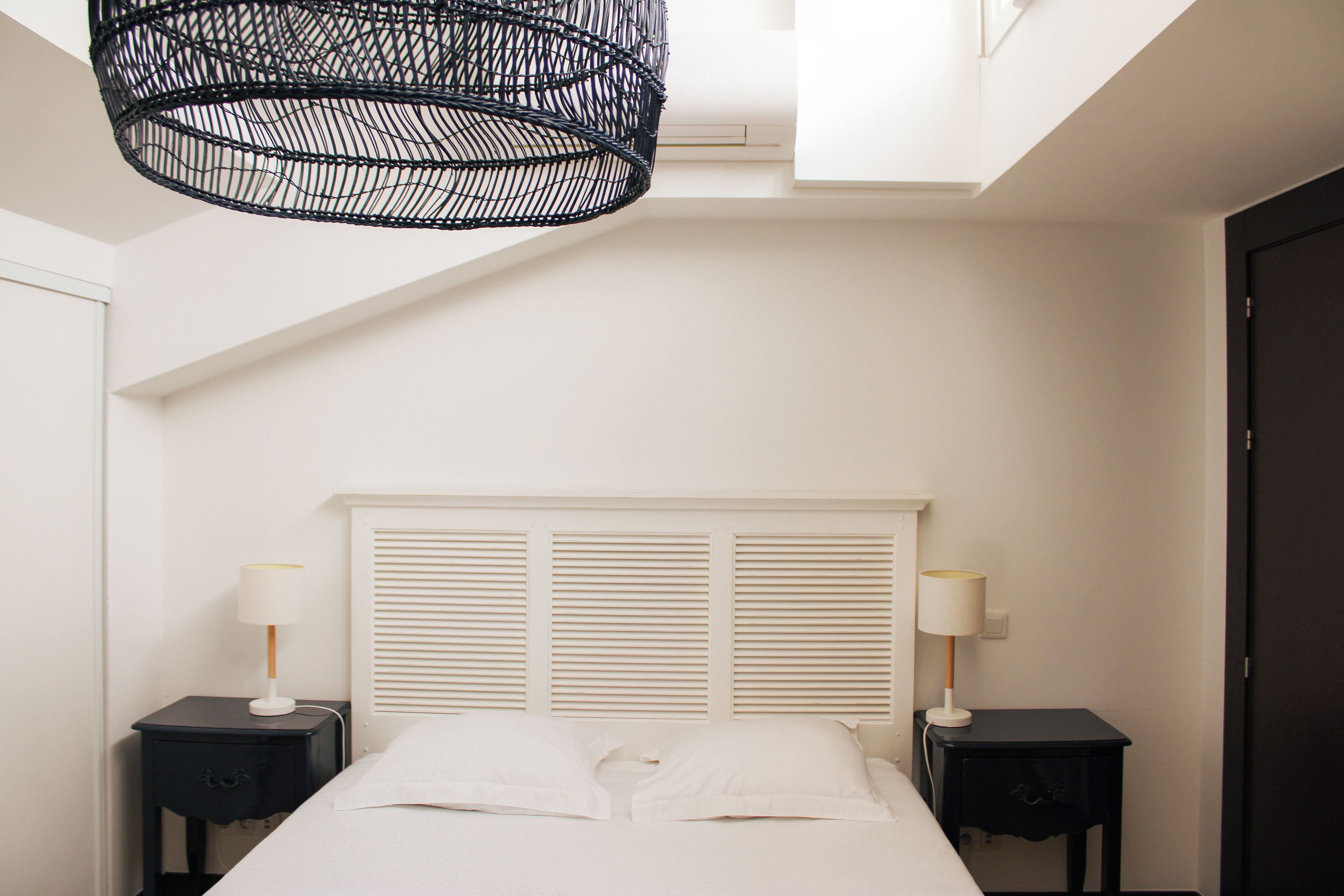 A bedroom with a bed, two bedside tables, a window with shutters, and a hanging light fixture.