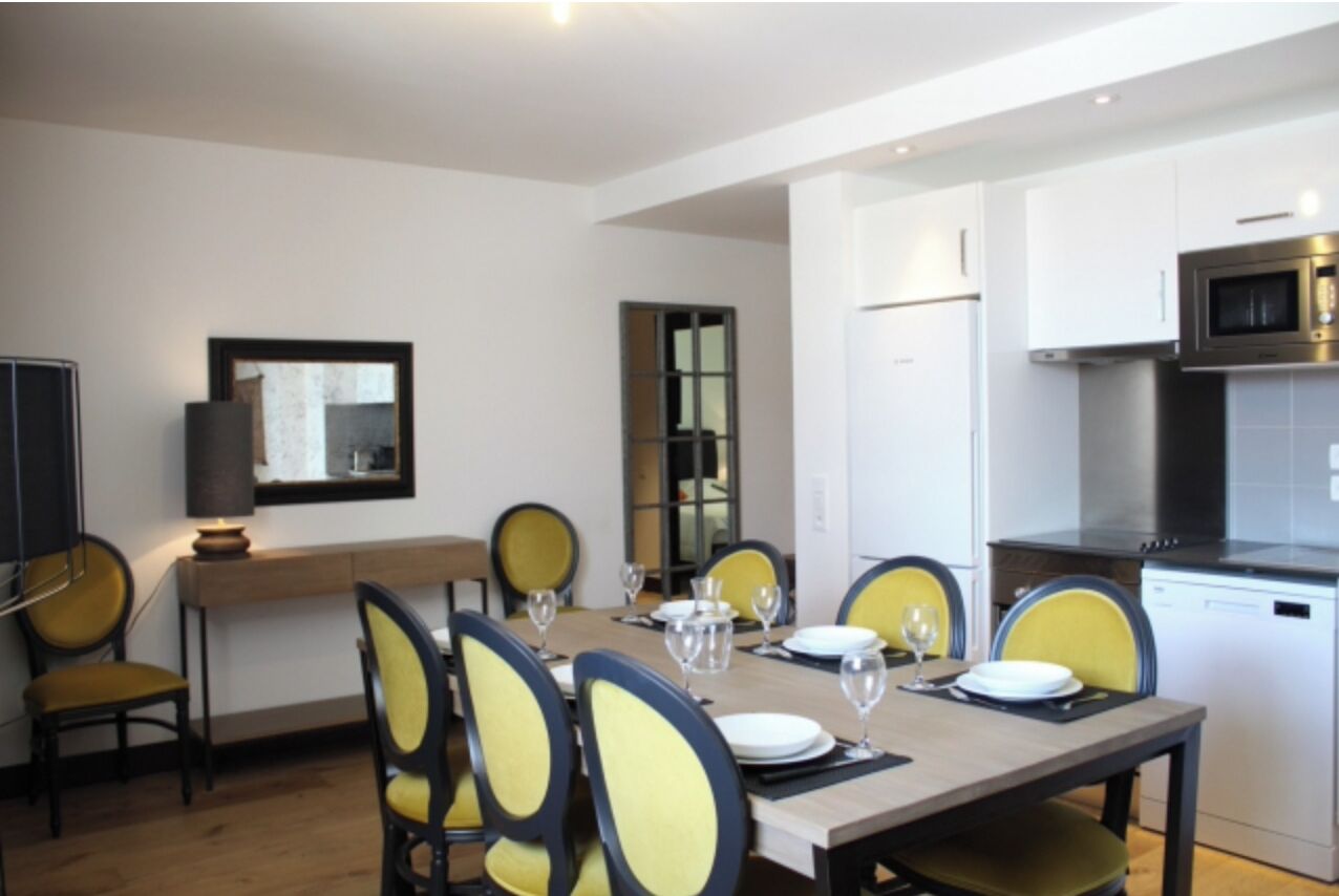 Un comedor con una mesa dispuesta para cuatro personas, rodeada de sillas amarillas. Se observa una zona de cocina con microondas y lavavajillas.