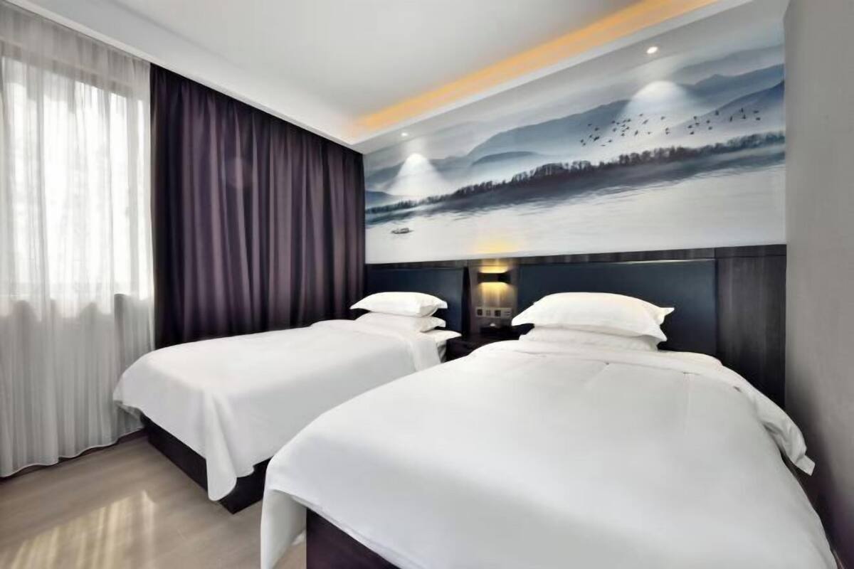 A hotel room with two beds, a large window with sheer curtains, and a mural of a landscape on the wall.