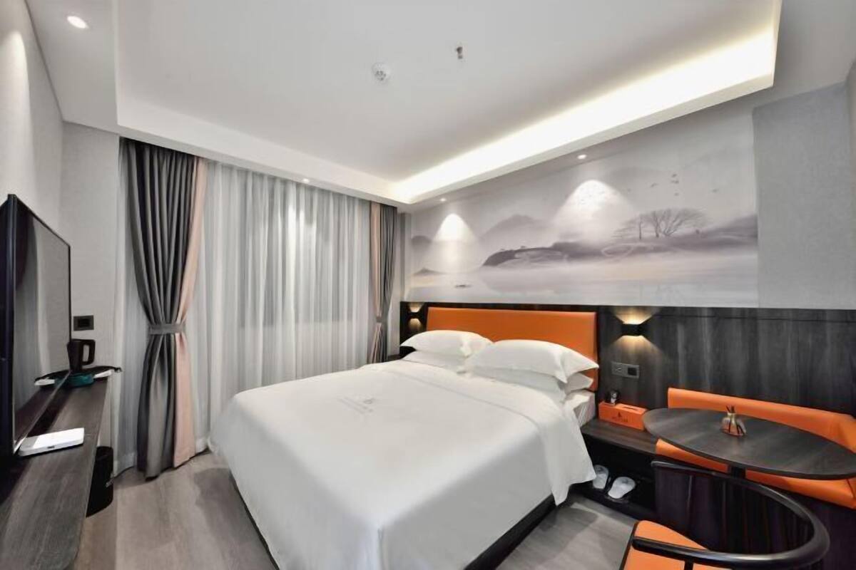 A modern hotel room with a large bed, a desk with a laptop, a small table, and orange seating.
