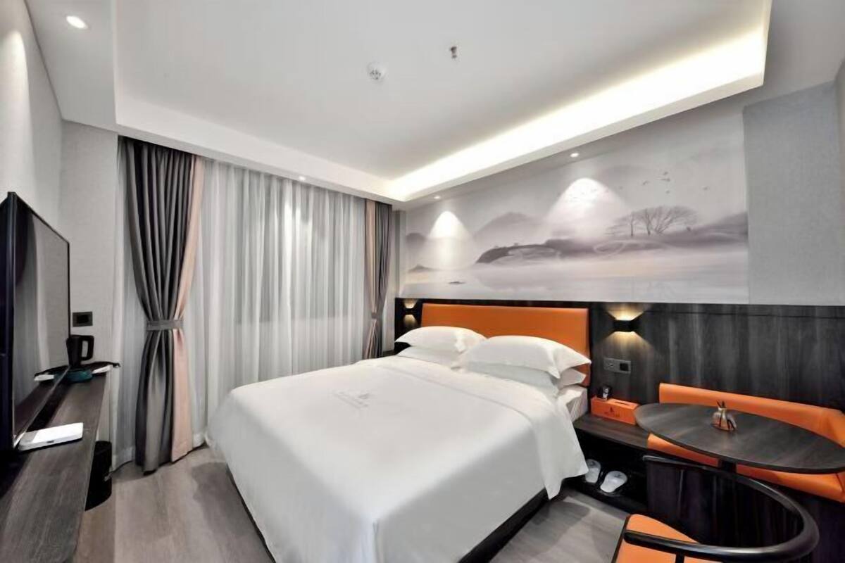 A modern hotel room with a large bed, a desk with a laptop, a small round table, and a seating area with orange cushions.