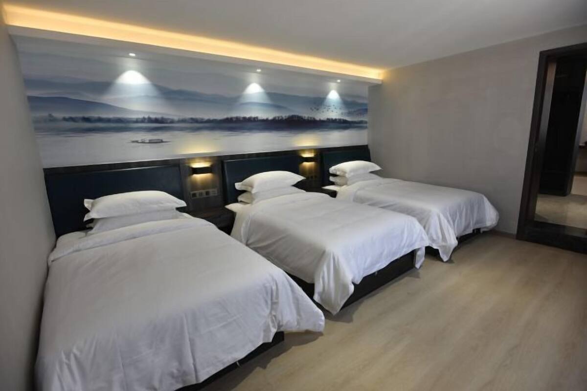 A hotel room with three single beds, a large wall painting of a lake, and a wooden floor.