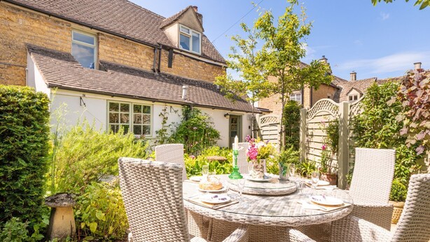 A garden patio with wicker furniture and a table set for dining, surrounded by lush greenery and a stone building. - Characterful holiday cottage in the heart of Bourton-on-the-Water - Pear Tree Cottage, Bourton (Cheltenham)