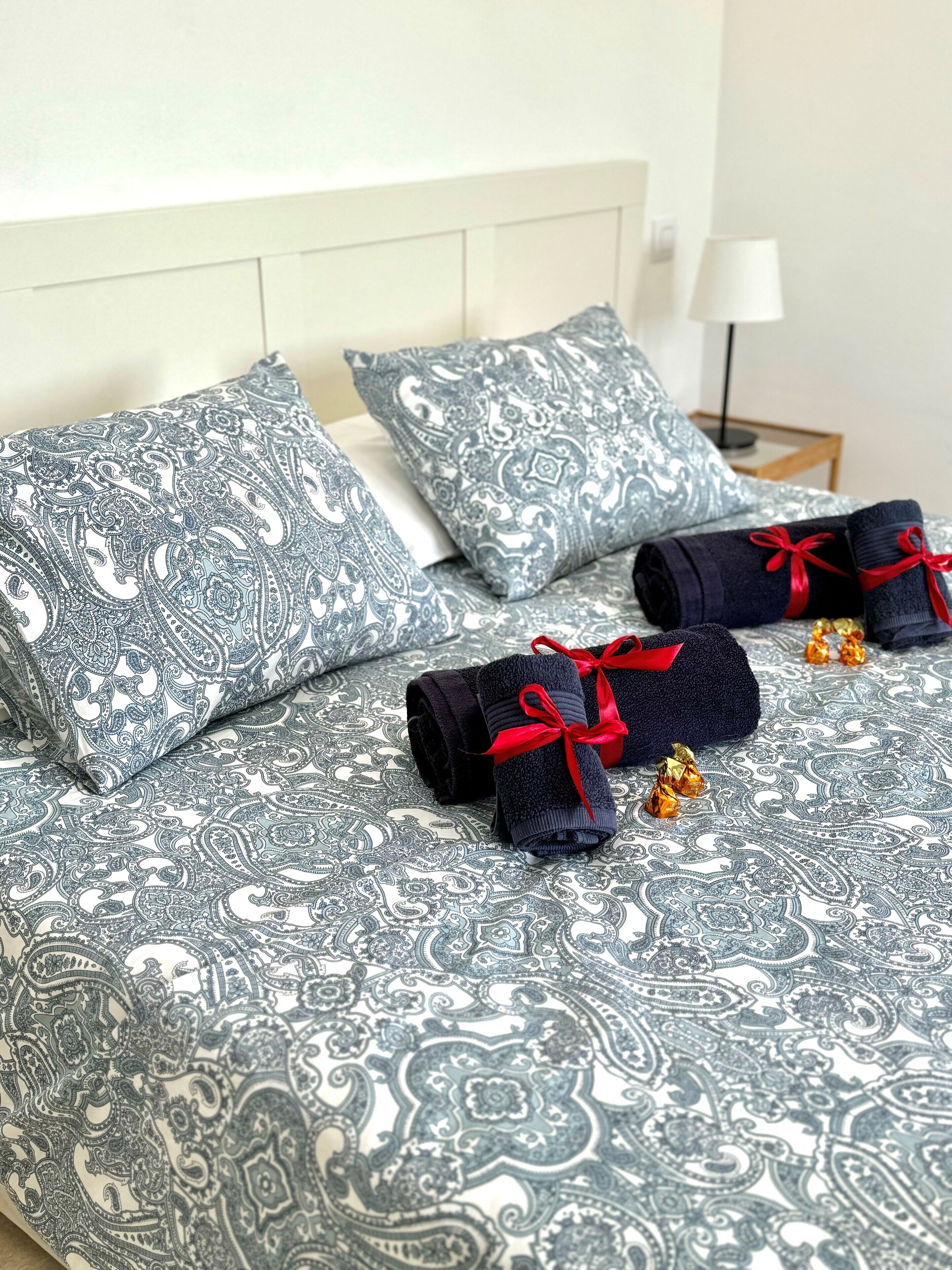 Een netjes opgemaakt bed met behang, twee opgerolde handdoeken met rode linten, en een bedlamp.