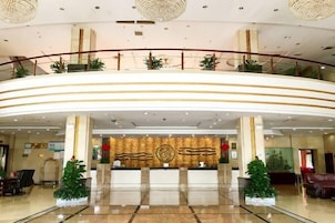 Un vestíbulo de hotel con un mostrador de recepción curvo, una pared decorativa con un emblema circular y una barandilla de cristal que da al vestíbulo.