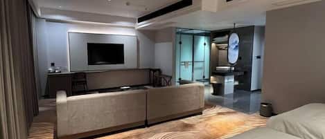 A modern hotel room with a sofa, a flat-screen TV, a dining area, and a bathroom with a mirror.