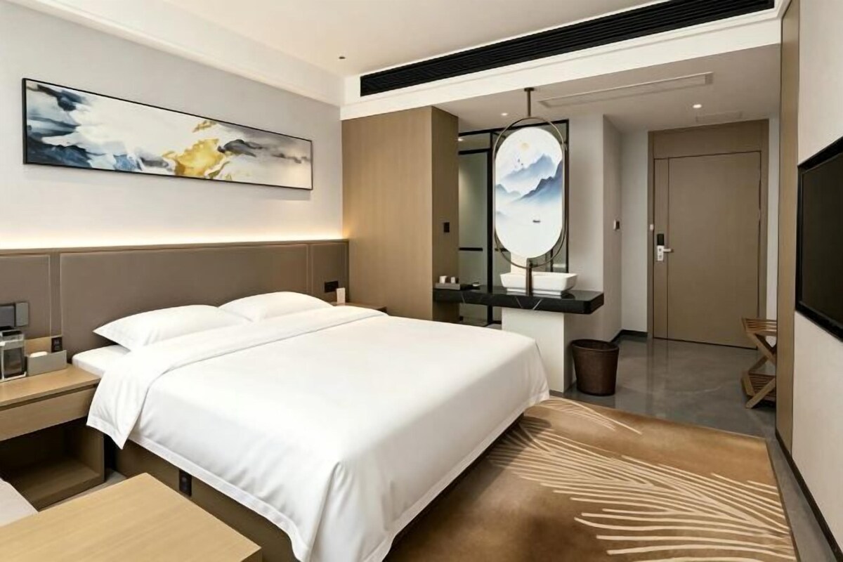 Une chambre d’hôtel moderne avec un grand lit, des tables de chevet, un téléviseur à écran plat, un grand miroir et un tableau au mur.
