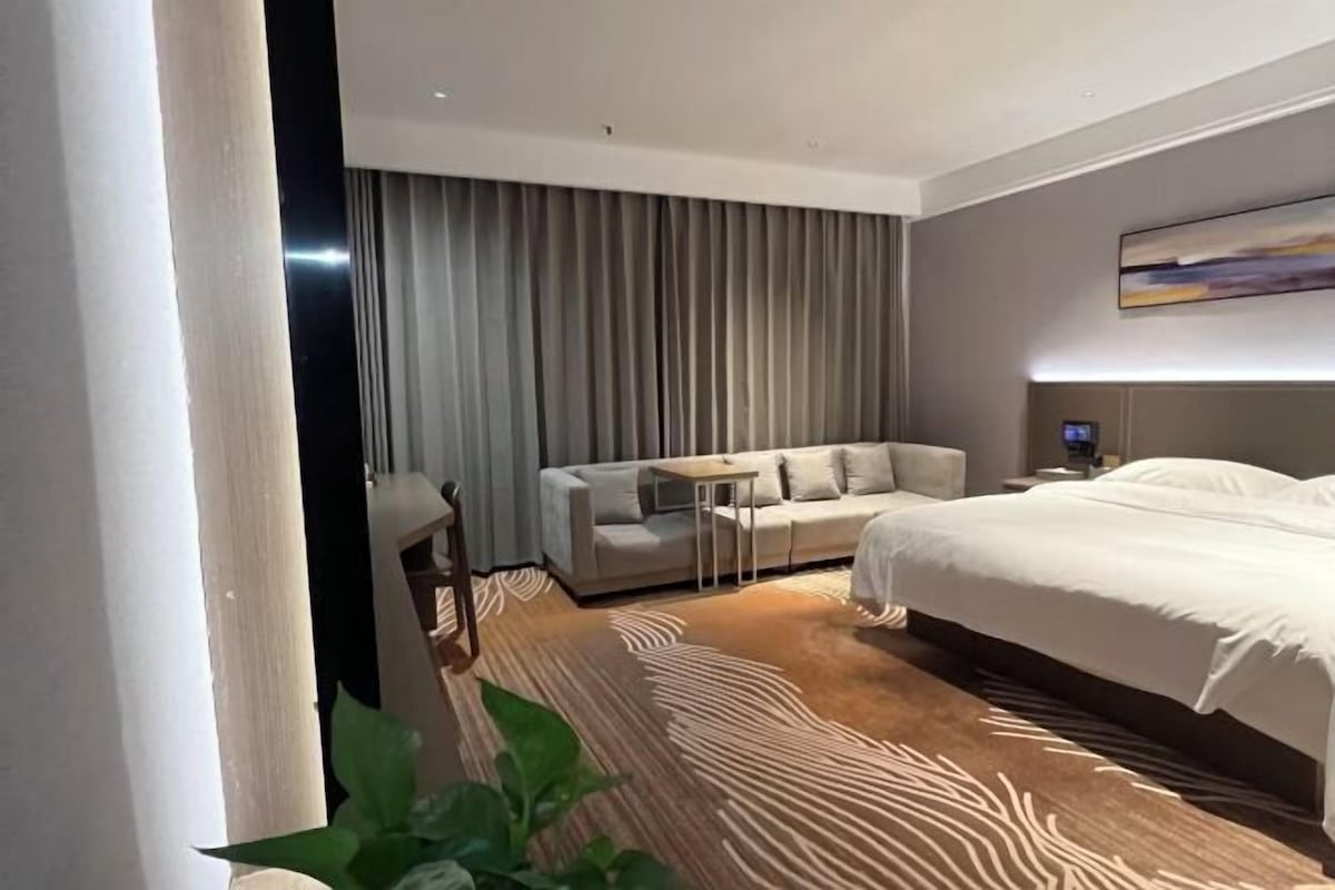 Moderni hotellihuone, jossa on suuri sänky, sohva, pieni pöytä ja suuri ikkuna, jossa on verhot.