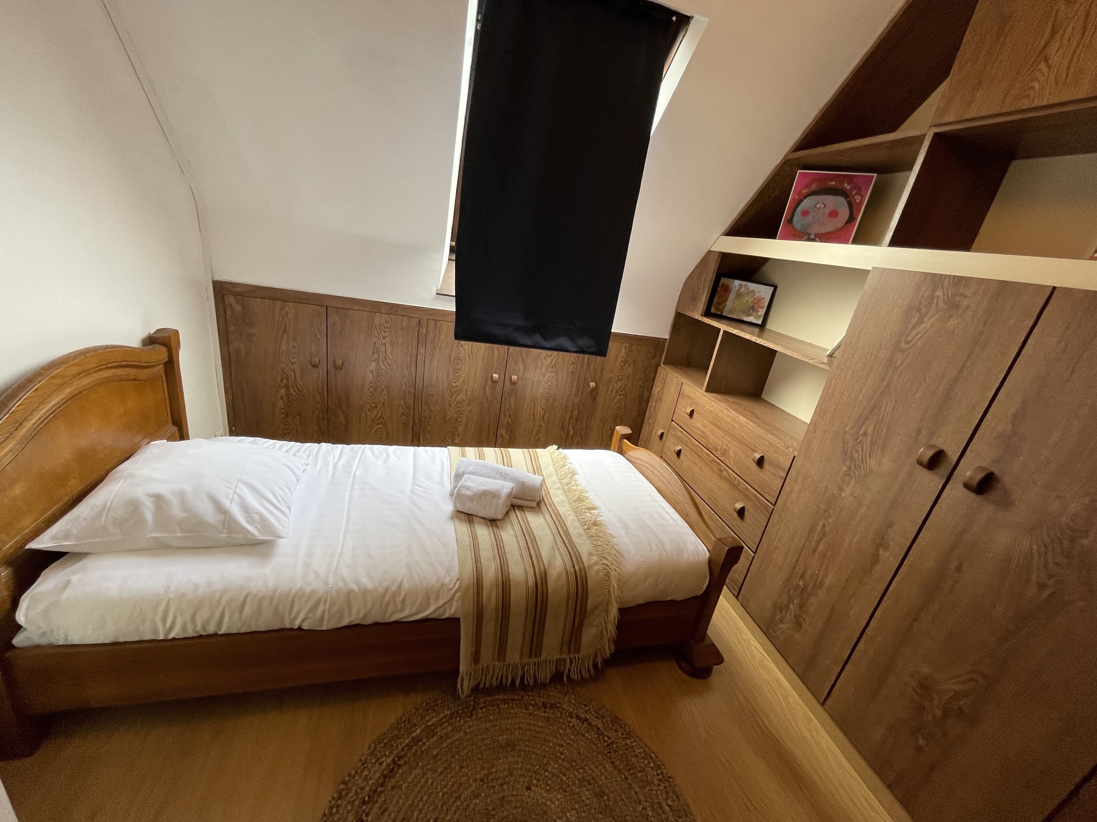 Ein Schlafzimmer mit einem hölzernen Bett, einem kleinen Fenster, einem Holzschrank und einem Regal mit einem Bild.
