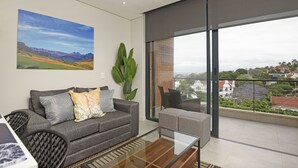 A modern living room with a grey sofa, a glass coffee table, and a large window offering a view of the cityscape.