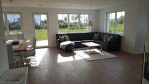 A spacious living room with a dark-colored sofa, a wooden coffee table, and a dining area with a table and chairs. Large windows provide ample natural light and views of a green outdoor area. - New, exclusive vacation home close to the beach with sauna, 3 bedrooms, fireplace, WLAN, garage (Dranske-Lancken)