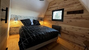 A cozy attic bedroom with a sloped ceiling, a bed with a black and white patterned bedspread, a stone accent wall, and a small window with a view of trees. - Chalet du Fayard in Belfahy, private Jacuzzi and view of the Vosges mountains (Belfahy)