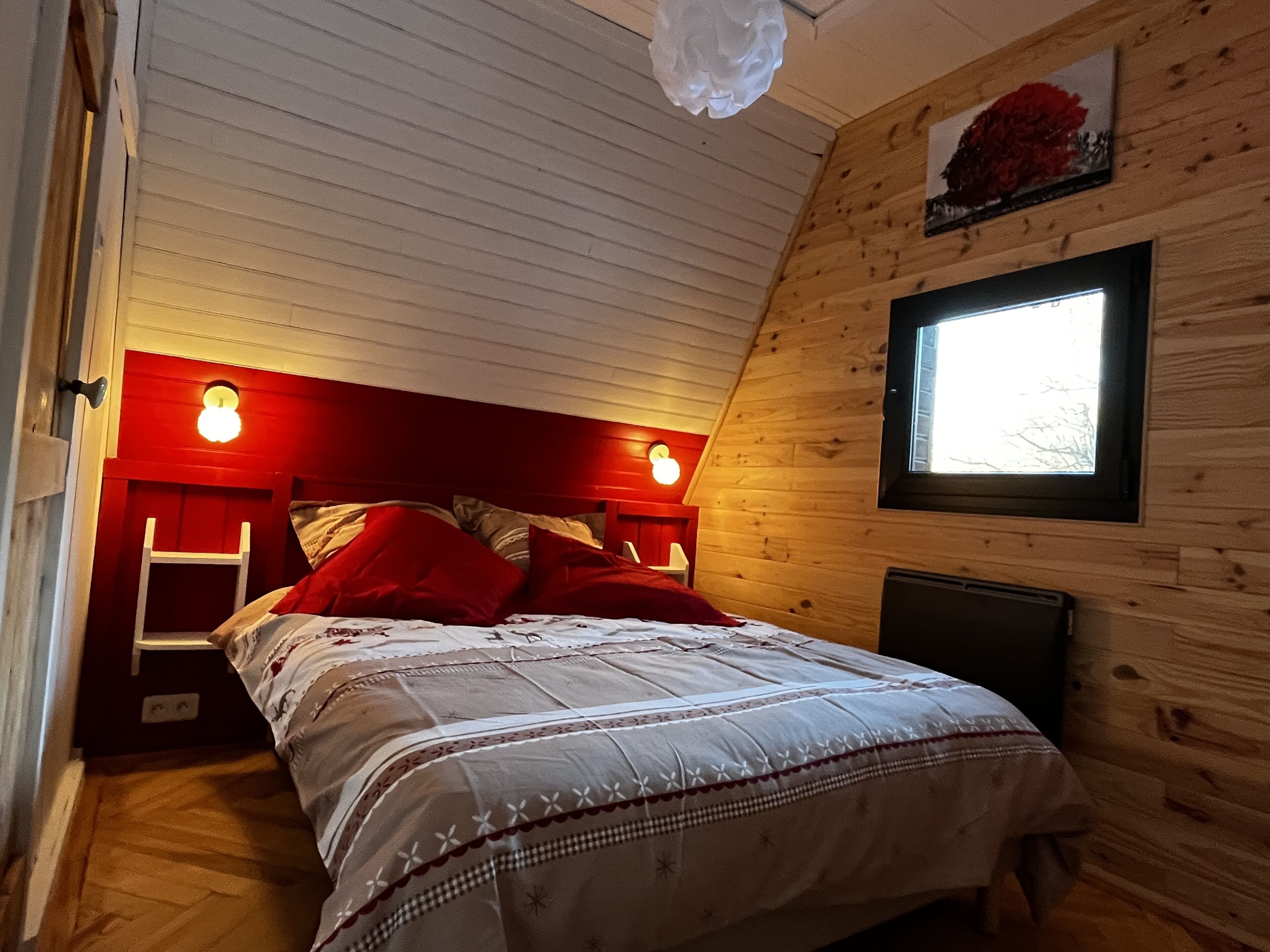 Ein gemütliches Schlafzimmer mit holzgetäfelter Schräge, einem Bett mit rot-weißer Bettwäsche, einer Leiter und einem Fenster mit Aussicht.