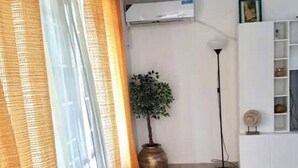 Ein Wohnzimmer mit großem Fenster, Klimaanlage, Stehlampe, Topfpflanze und einem weißen Regal.