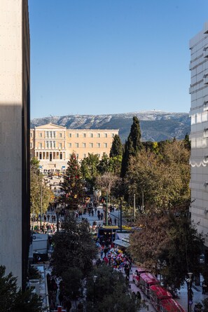 Une vue d’une place publique, avec un grand bâtiment en arrière-plan, des arbres et un ciel dégagé.