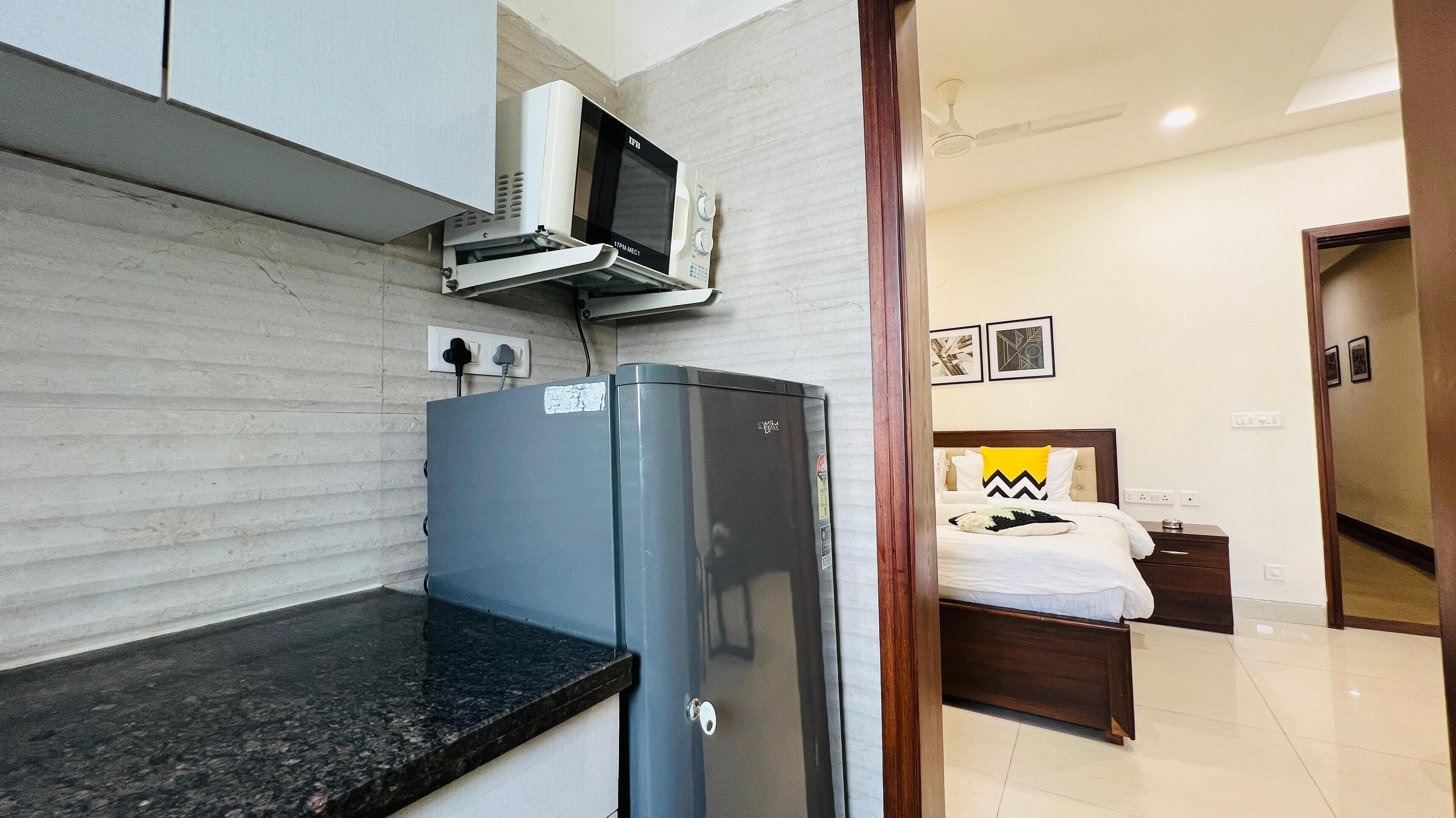 A compact kitchenette with a microwave and a refrigerator, a bed with a wooden headboard and decorative pillows, and a hallway leading to a bathroom.