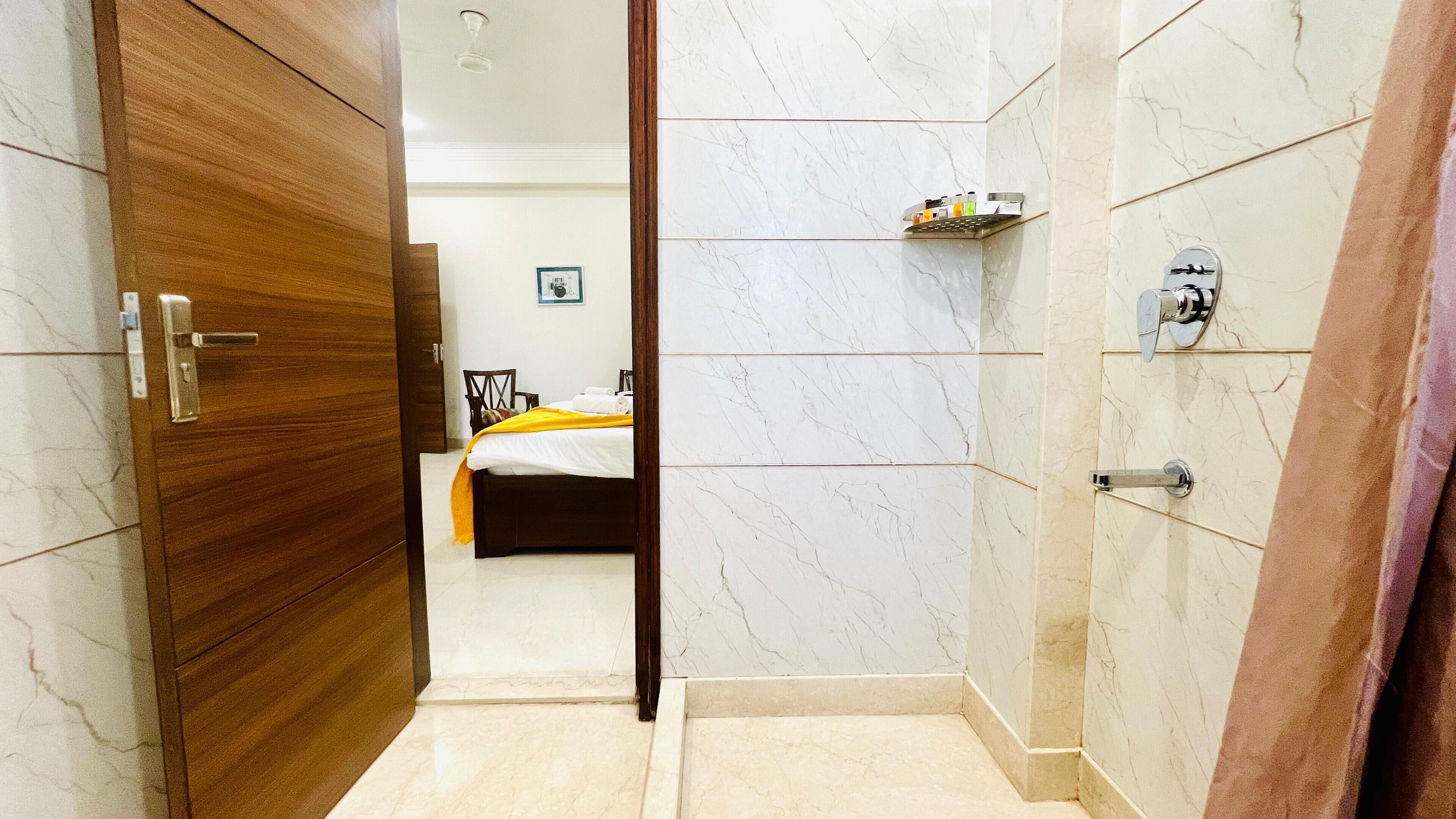 A hotel room with a wooden door, a bed with a white and yellow bedspread, a small shelf with toiletries, and a bathroom with marble walls and a shower.