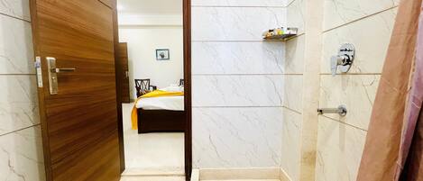 A hotel room with a wooden door, a bed with a white and yellow bedspread, a small shelf with toiletries, and a bathroom with marble walls and a shower.