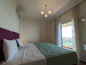 A hotel room with a bed, a chandelier, an air conditioning unit, a balcony with a view, and a window with curtains.