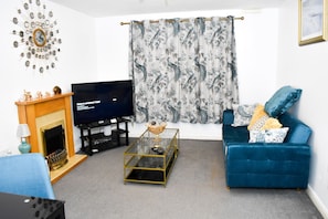 A living room with a TV, a fireplace, a blue sofa, and a floral curtain. - Lovely 2-bed Apartment in West Bromwich (West Bromwich)