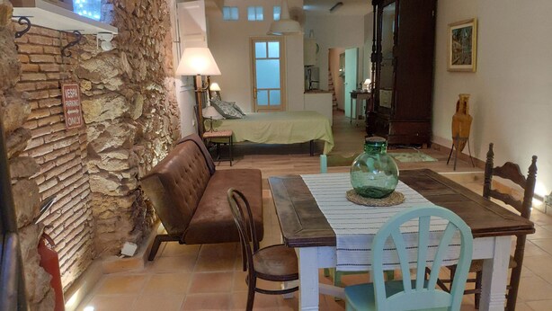 Un comedor acogedor con pared de piedra, un sofá de cuero marrón, una mesa de madera con un jarrón verde y una cama al fondo.
