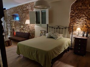 Un dormitorio con pared de piedra, cama con colcha verde a cuadros, mesita de noche de madera con lámpara y chimenea de piedra con luz azul.