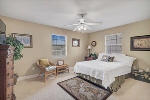 Une chambre à coucher comprenant un lit, un ventilateur de plafond, une commode, une chaise, une fenêtre avec des stores et un tapis.