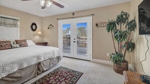 Un dormitorio con cama, ventilador de techo, ventana con persianas, puerta a un balcón y una planta en maceta.