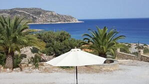 Un parasol blanc dans un jardin planté de palmiers, avec une vue sur la mer et une colline rocheuse en arrière-plan.