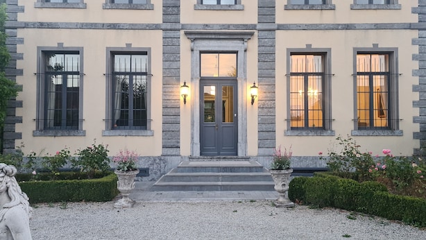 A stately building with a symmetrical facade, featuring a central entrance with a dark door, flanked by two windows with light fixtures. - Le Château des Comtesses (Montignies-sur-Roc)
