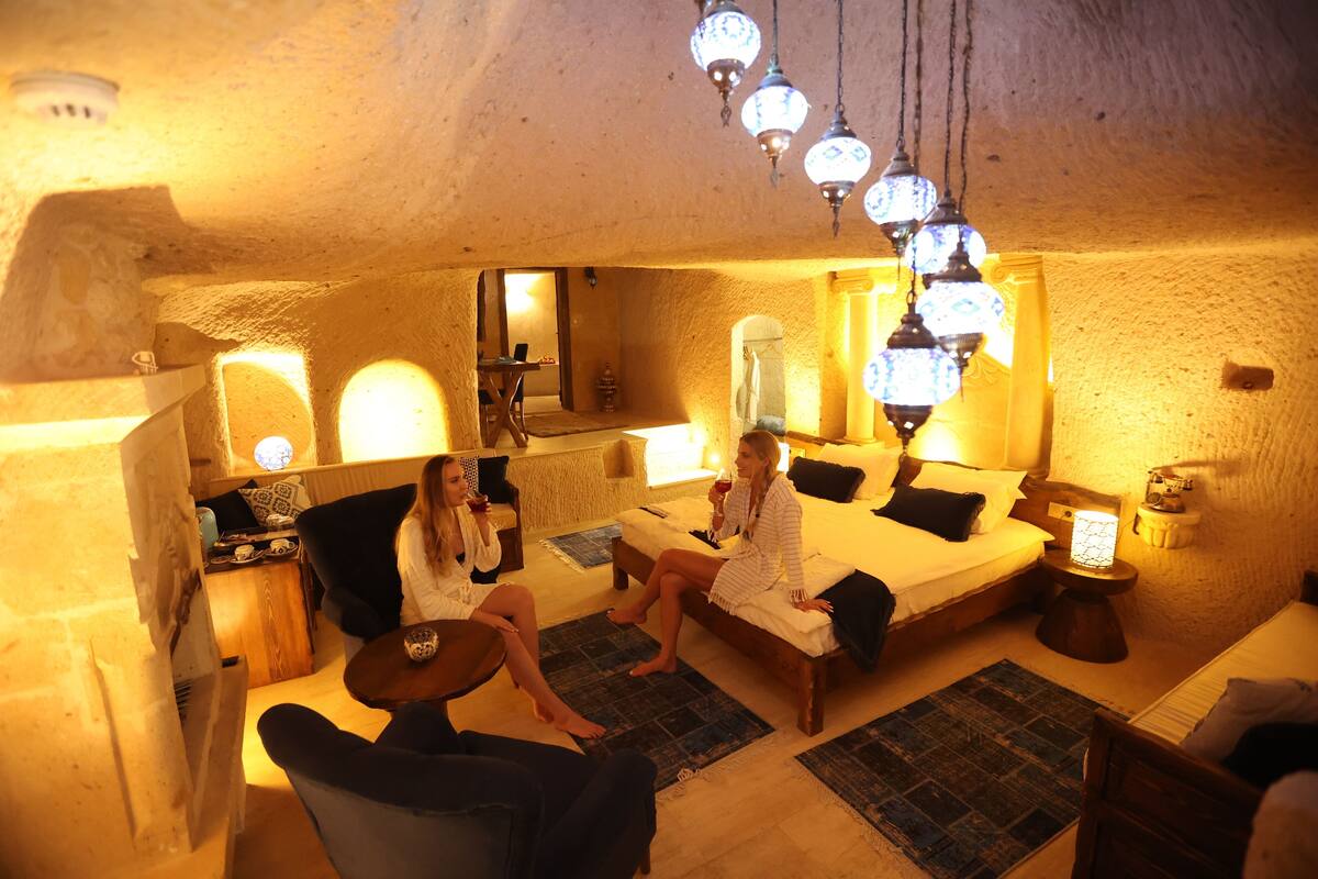 Quarto aconchegante e bem iluminado, com uma cama grande, uma mesinha e uma cadeira. Há luminárias pendentes e um espelho na parede.