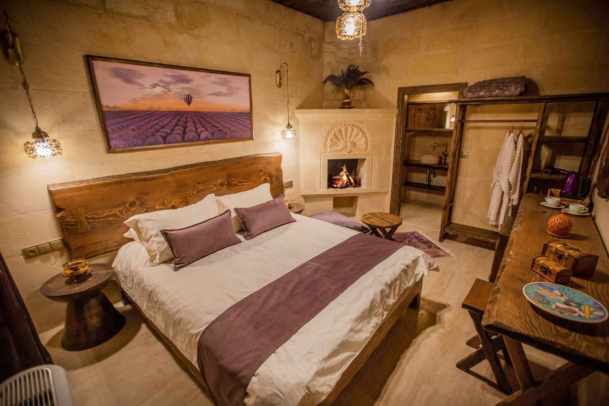 Um quarto aconchegante com uma cama grande, uma lareira, um aparador de madeira e uma pintura de um pôr do sol sobre um campo.