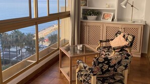 Une pièce dotée d’une grande fenêtre donnant sur une plage, d’une table en verre, d’un fauteuil à motifs floraux et d’une étagère ornée d’objets décoratifs.