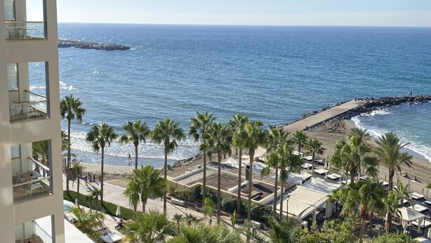Un hôtel en bord de mer, doté de balcons offrant une vue sur l’océan, des palmiers et une digue rocheuse.