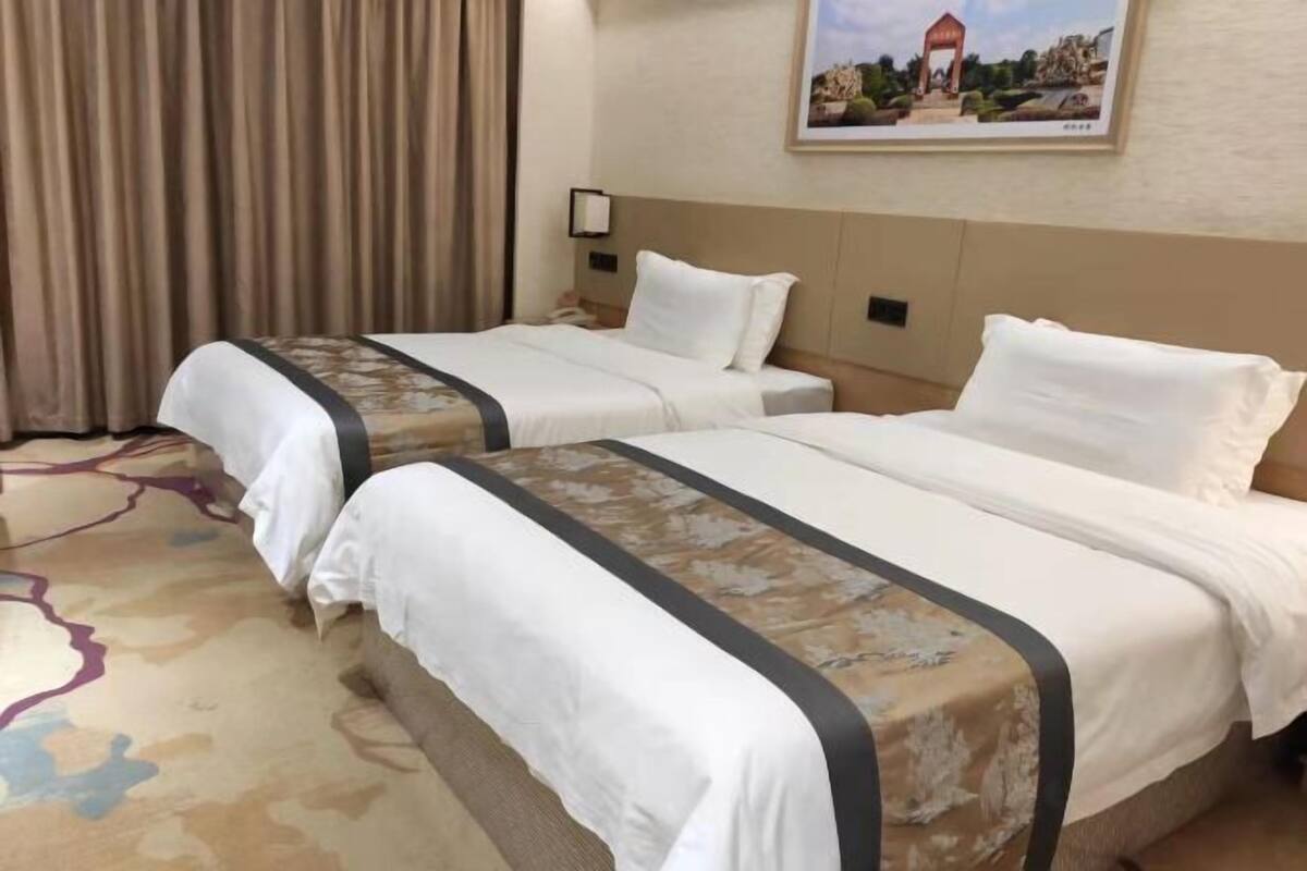 Een hotelkamer met twee bedden, een groot raam met gordijnen, een lijst met een afbeelding aan de muur en een vloerbedekking.
