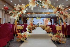 Un salón de banquetes con mesas dispuestas en filas, un arco floral en la entrada y una zona de escenario al fondo.